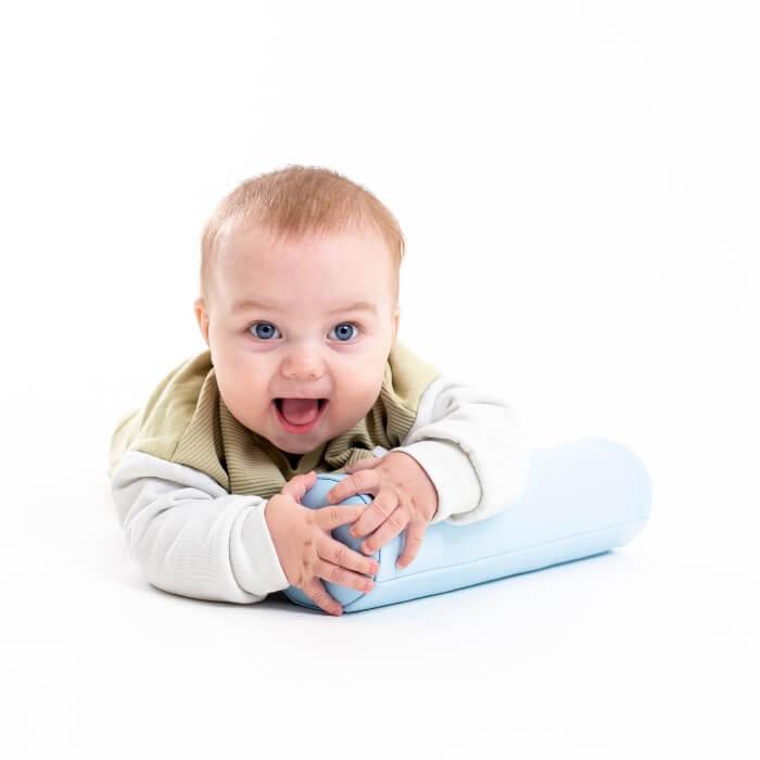 Excited baby with blue eyes, wearing light-colored clothes, hugging a cylinder-shaped soft play tummy roller