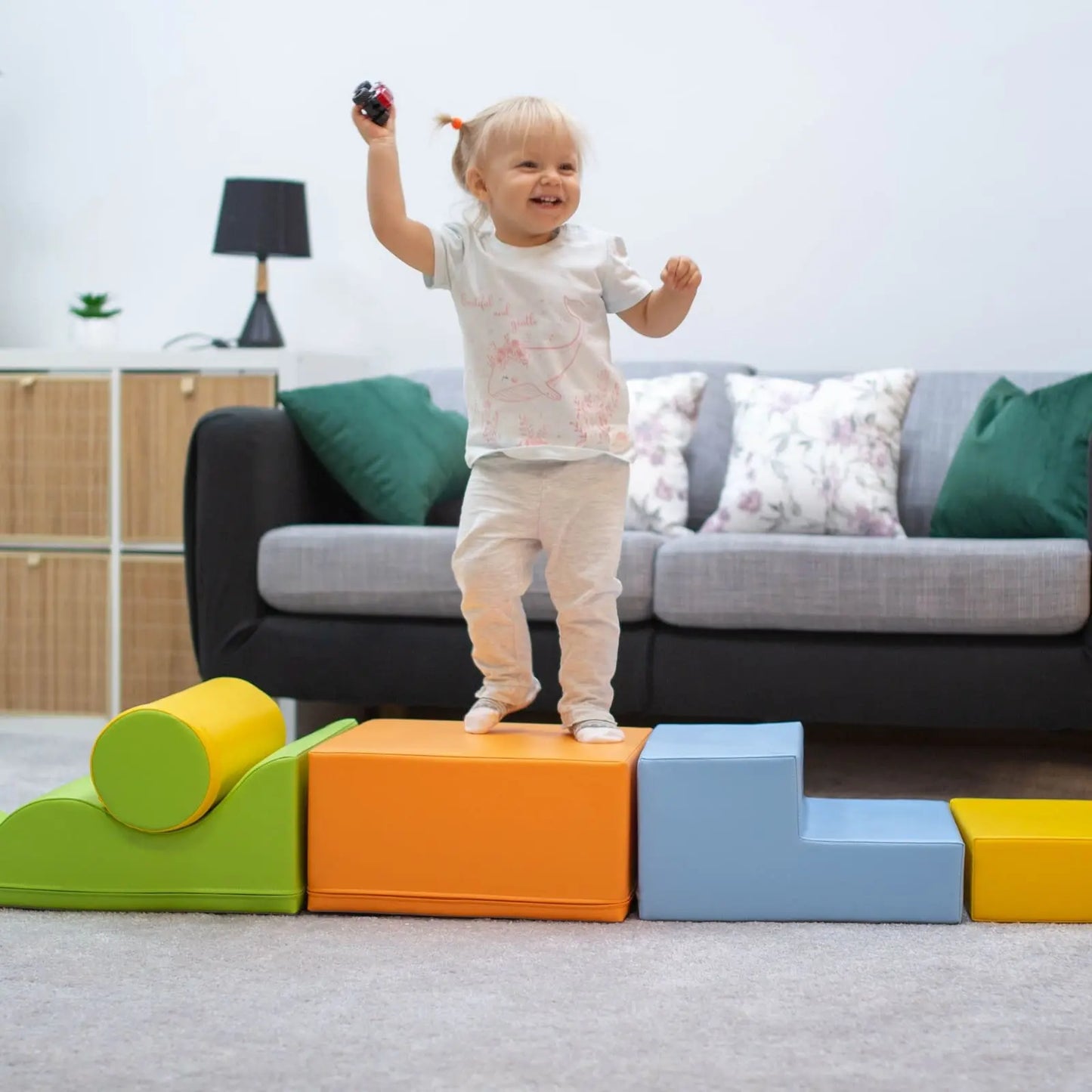 Toddler girl laughing on a colorful soft play set arranged from various shapes like squares and steps in the living room