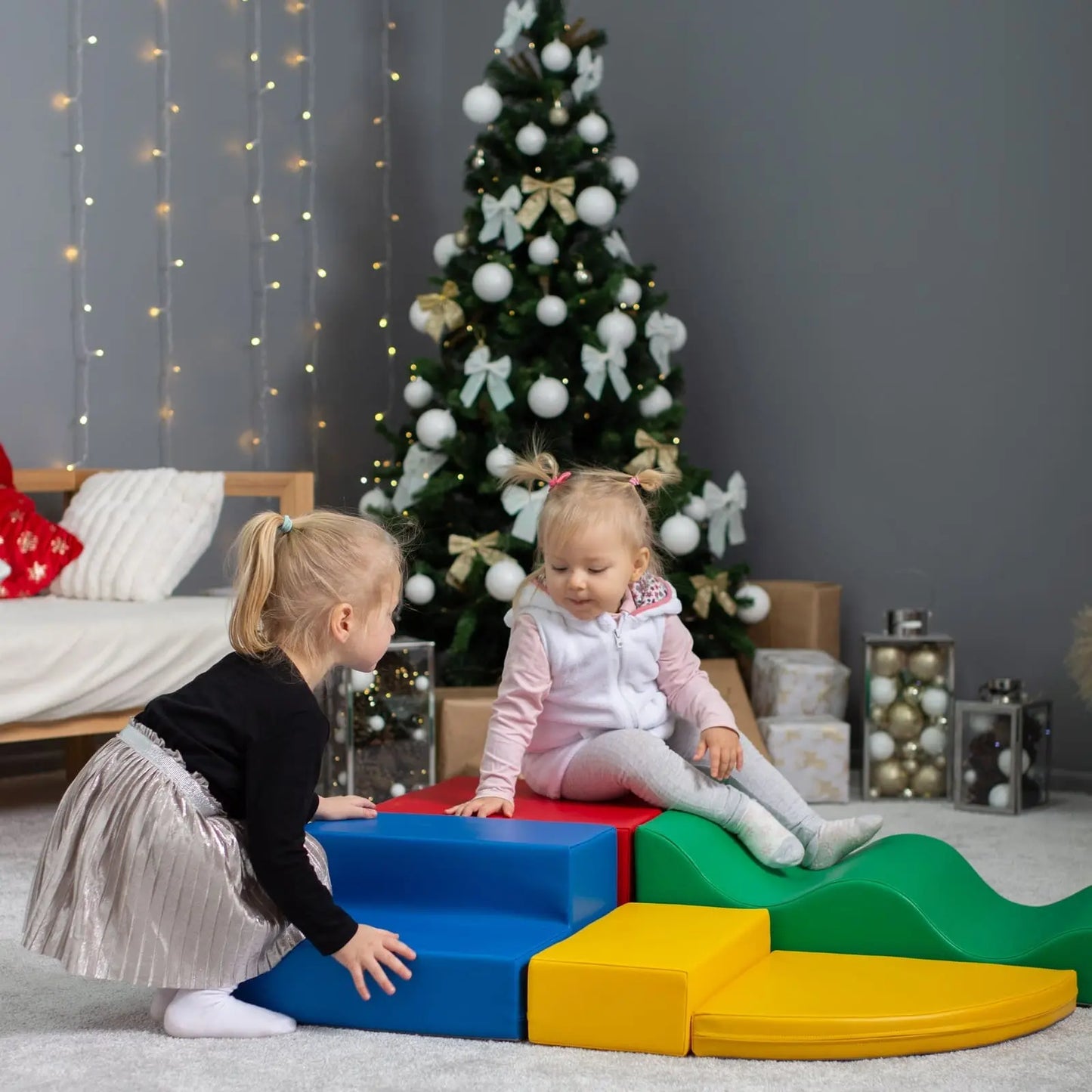 Two young girls playing on a colorful soft play set in front of a Christmas tree, consisting of squares, steps, and other shapes