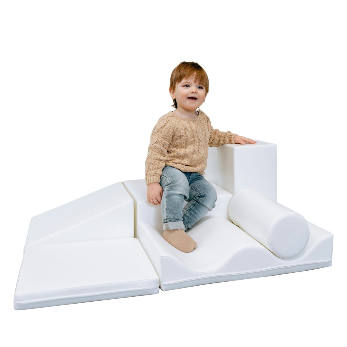 Two-year-old boy patiently sitting on a white soft play set made from different shapes, looking curious and happy