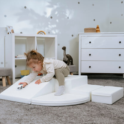 There is a toddler playing with toy car on white soft play equipment which is arranged from various shapes like squares and steps