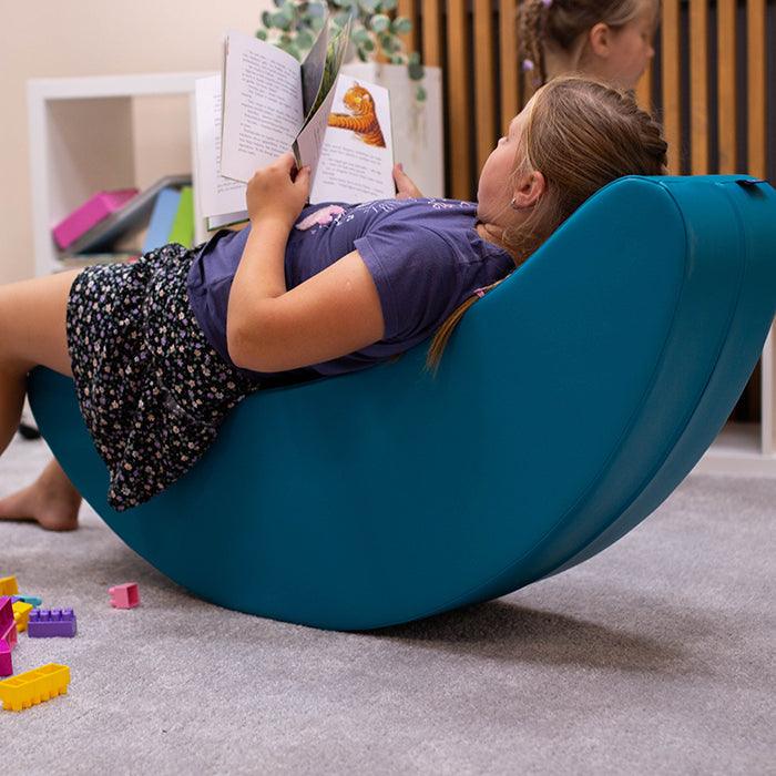 Girl reading a book laying on her blue banana shaped rocker toy