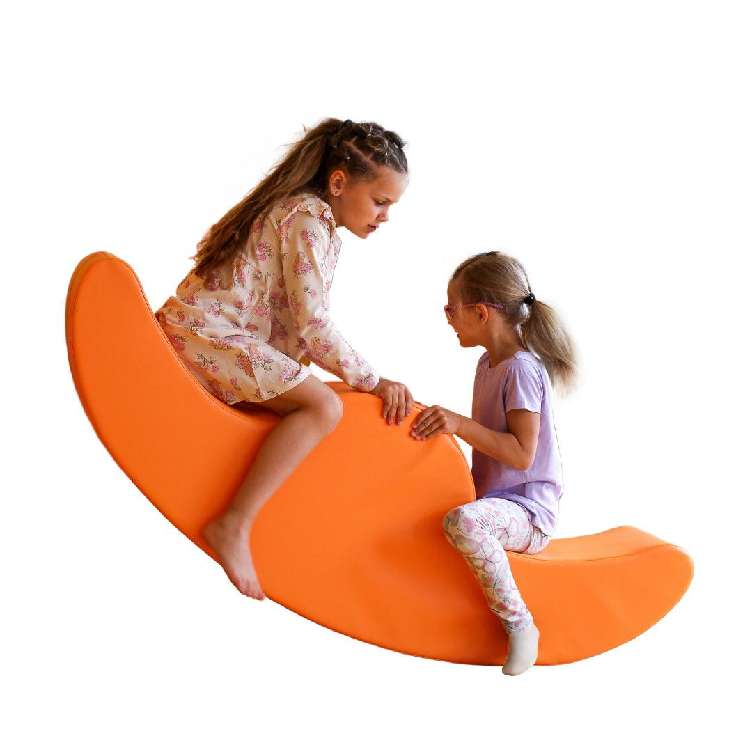 Two kids rocking on an orange, two-seated soft play rocker which has a round shape