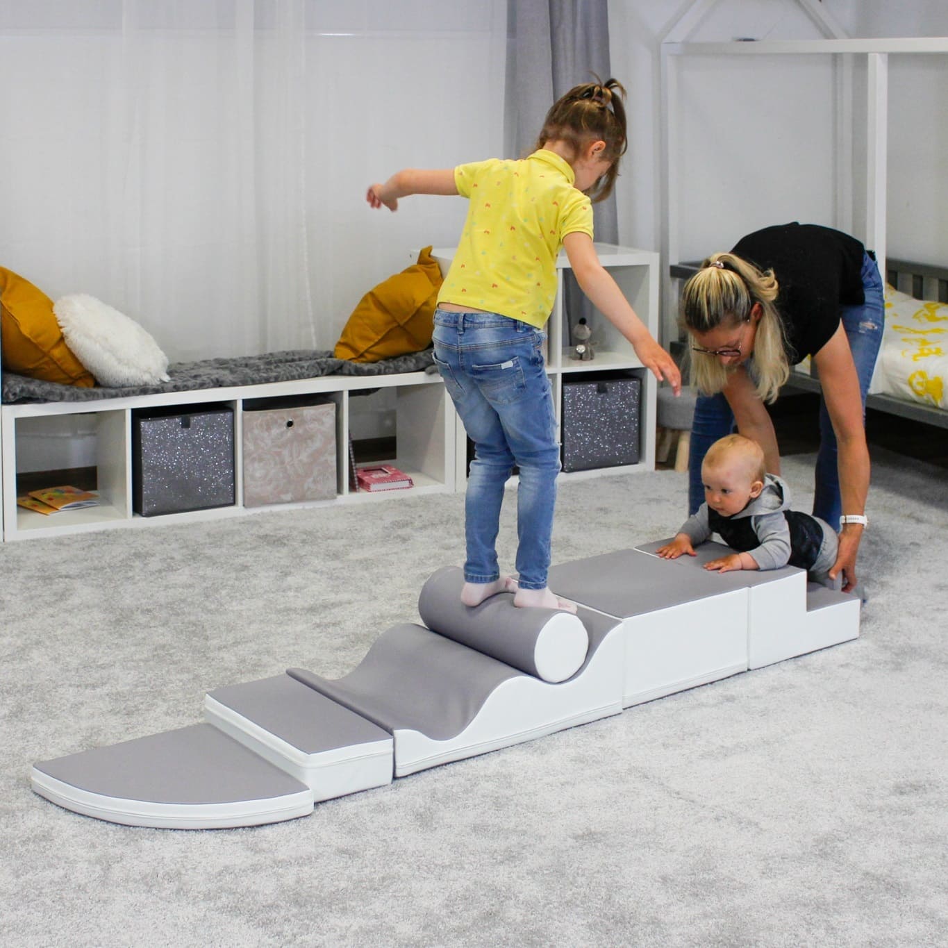 Girl balances and a baby crawls on gray-white soft play set in a room, made from various shapes like a square, wedge, cylinder