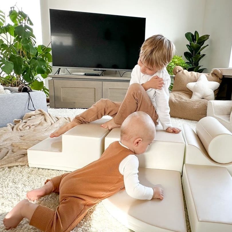 Two children playing on a beige soft play set. Older child sits, younger crawls on modular foam blocks in living room setting